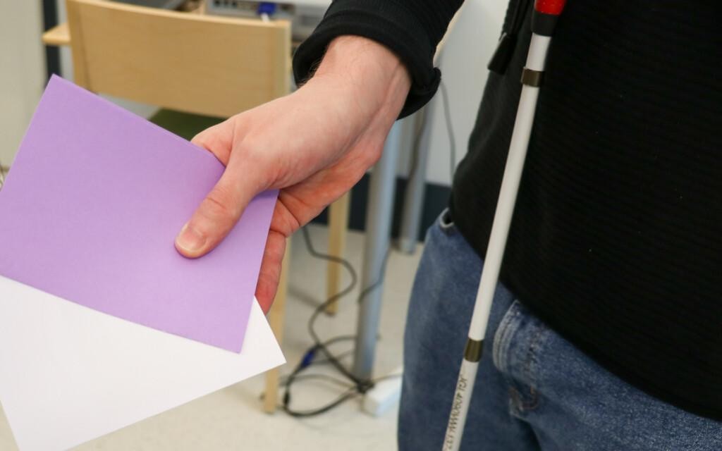 Lähikuva kädestä, jossa on kirjekuori ja lappu. Kuvan oikeassa reunassa näkyy valkoinen keppi.