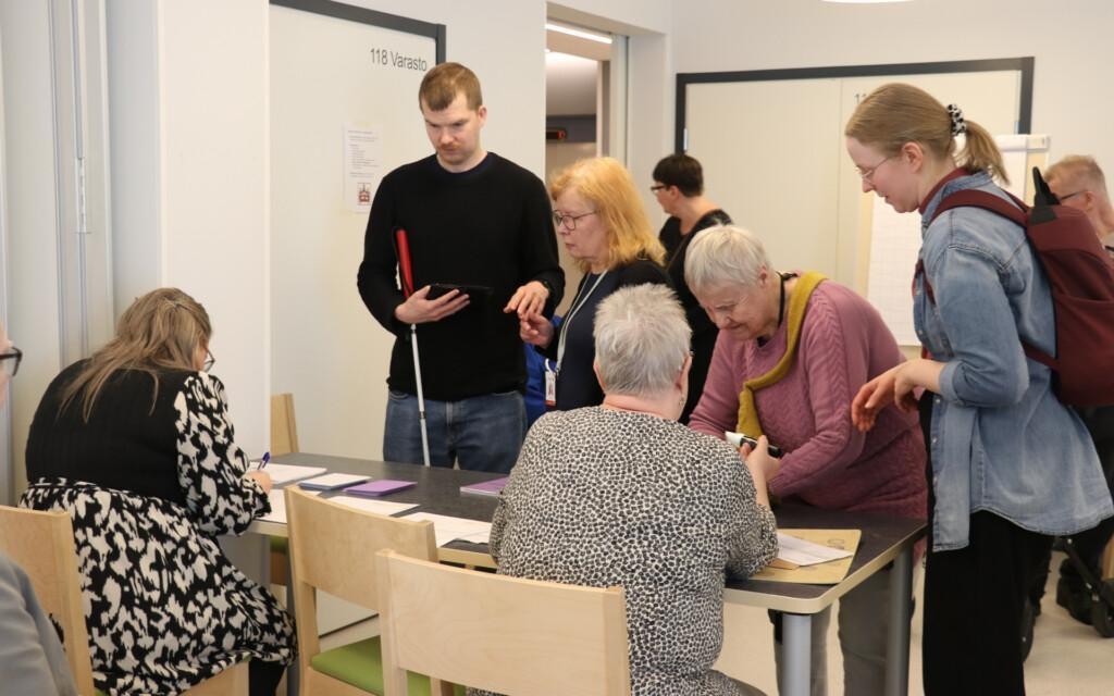 Henkilöitä äänestystilanteessa. Kaksi henkilöä istuu pöydän ääressä, neljä seisoo pöydän edessä. Yhdellä henkilöllä on kädessään valkoinen keppi